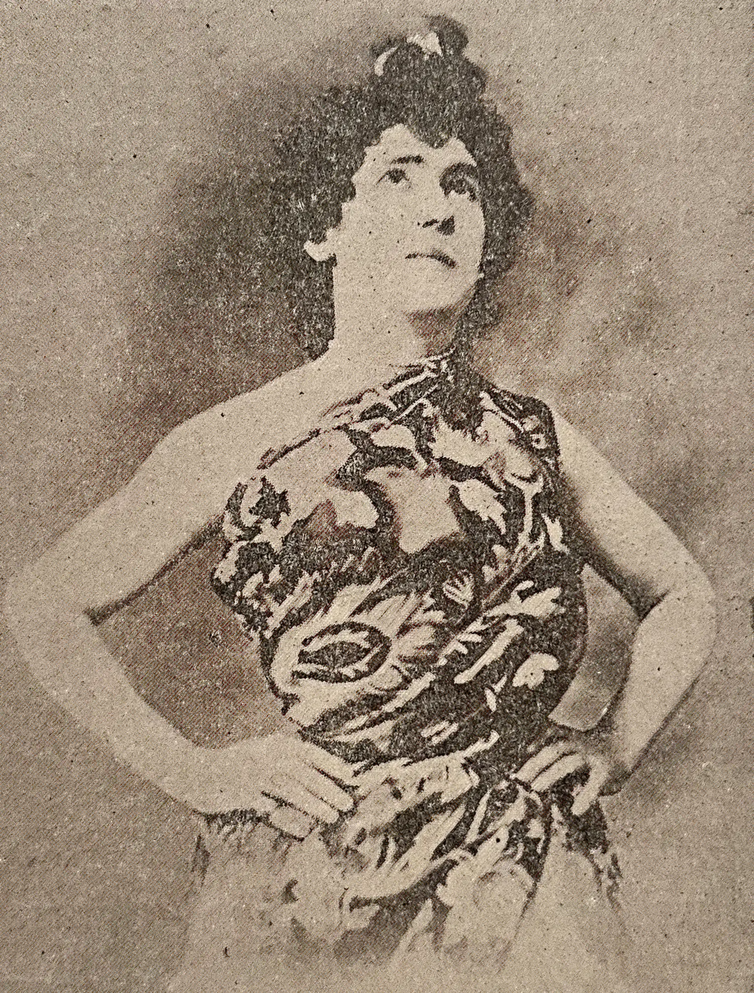 A grainy old photograph of a feminine-presenting person with curly hair wearing an intricately patterned garment. The person stands with their hands on their hips, contemplatively looking up.